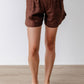 Woman wearing a brown linen shorts standing against a white wall.