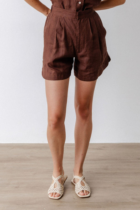 Woman wearing a brown linen shorts standing against a white wall.
